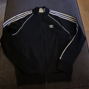 Svart träningsjacka från Adidas - Säljer en klassisk svart träningsjacka från Adidas med vita ränder längs ärmarna och logga på bröstet. Jackan har dragkedja och långa ärmar, perfekt för sportiga tillfällen.