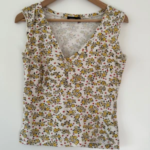 Blommig ärmlös topp från Lindex - Säljer en söt ärmlös topp från Lindex med ett blommigt mönster i gult, rött och svart på en vit bakgrund. Toppen har en v-ringad design och är perfekt för varma dagar. Passar bra till både jeans och kjol!