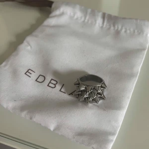 Silvrig ring med nitar från Edblad - Snygg och unik ring i silver från Edblad, dekorerad med coola nitar runtom. Perfekt för att ge din outfit en edgy touch. Kommer med en praktisk förvaringspåse.