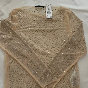 Beige mesh topp från Gina Tricot - Säljer en snygg beige topp från Gina Tricot. Toppen är dekorerad med diamanter över hela ytan och har långa ärmar. Den är helt ny med prislapp kvar. Är i st L men passar S.