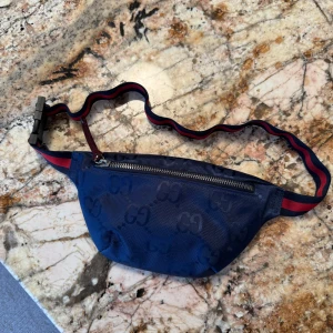 Blå magväska från Gucci - Säljer en snygg blå magväska från Gucci med det klassiska GG-mönstret. Väskan har en dragkedja och ett justerbart band i rött och blått. Perfekt för att bära dina essentials med stil. Inuti är den fodrad med rött tyg.