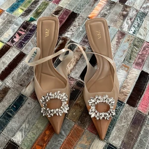 Beige klackskor med strassdetaljer från Zara - Eleganta beige klackskor från Zara med spetsig tå och glittrande strassdetaljer framtill. Skorna har en smal klack och transparenta remmar som ger en stilren look. Använda endast 1 gång 