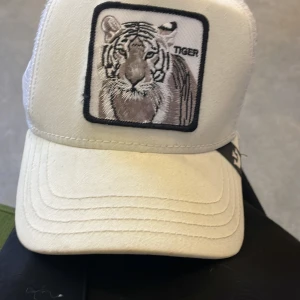 Vit keps med tigerbroderi - Snygg vit keps med ett broderat tigeremblem på framsidan. Kepsen har en justerbar baksida i meshmaterial för bra ventilation. Perfekt för en avslappnad stil.