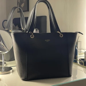 Svart handväska från Guess - Snygg svart handväska från Guess med elegant design. Väskan har guldfärgade detaljer och en praktisk dragkedja upptill. Perfekt för att bära dina dagliga nödvändigheter med stil. Rostig på guld detaljerna!