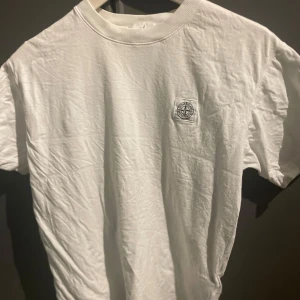 Vit t-shirt från Stone Island - Säljer en stilren vit t-shirt från Stone Island med deras ikoniska logga broderad på bröstet. Perfekt för en avslappnad och trendig look. T-shirten har en klassisk passform och är tillverkad i mjukt bomullsmaterial.