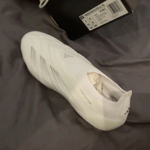 Vita fotbollsskor Adidas Predator Elite - Snygga vita fotbollsskor från Adidas Predator-serien. Skorna har en modern design med diskreta detaljer och är perfekta för spel på gräs. De är lätta och bekväma, vilket ger bra kontroll och grepp på planen. SKRIV jätte gärna om ni är intresserade? Andvönda 3 gånger endast