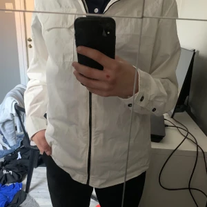Vit overshirt från C.P. Company - Säljer en stilren vit overshirt från C.P. Company. Overshirten har en dragkedja framtill och långa ärmar. Den är prydd med en rund svart detalj på ärmen med märkesloggan. Perfekt för en sommarkväll eller vårdag.