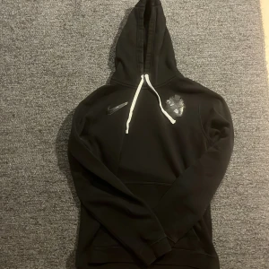 Svart AIK hoodie från Nike - Säljer en svart AIK hoodie från Nike med vit dragsko och ett emblem på bröstet. Perfekt för en sportig look eller avslappnad stil. Hoodien har långa ärmar och en klassisk passform.