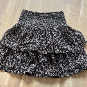 Kort volangkjol - En kort blommig volangkjol. 100% polyester. Svart kjol med små lila blommor på. Från Kappahl i stl 146-152. Använd ett fåtal gånger