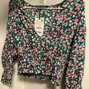 Kort blommig blus/topp - Oanvänd kort blus/topp med blommigt mönster i grönt, rosa och svart. Den är v-ringad och har lite puff i axlarna. Det är resår bak i midjan och knappar längst ut i ärmarna. Storlek L men den är liten i storlek och därför upplagd som M. Köpt för 359kr.