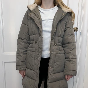 Zara vinterjacka - Skön täckjacka från Zara. Supermjukt foder innanför luvan. Modellen är rymlig och går ner en bit över låren. Knappt använd, bara enstaka gånger. Guldiga detaljer och dragsko vid midjan.  (Passar mig som är 150 cm lång.)