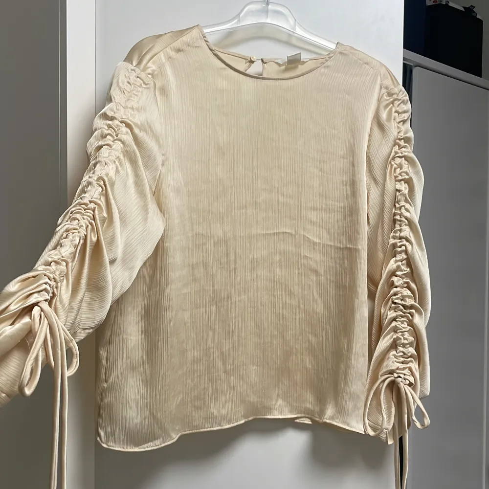 Helt ny satin blus från H&M i storlek 38. Armarna har en fin modell där man kan justera själv hur ”tajt” man vill ha dem. Den har en skinande beige färg. Utan prislapp. . Puserot.