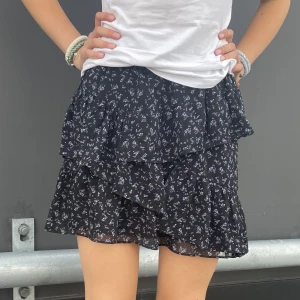 Svart volangkjol - Svart volangkjol med små lila och rosa blommor som mönster. Ganska lång. Polyester