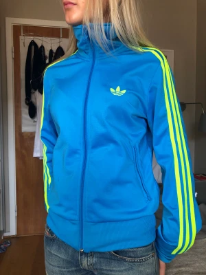 Adidasjacka - Väldigt trendig adidasjacka i färgen blå med neongula detaljer😍 köpt second hand men endast använd 1 gång av mig! Storlek 38 men passar även mindre och större beroende på hur man vill att den ska sitta!