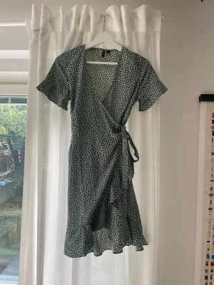 Prickig omlottklänning - Gröngrå knälång klänning med prickar. Omlott-modell med knyte, volanger längs armar och nederkant. Använd vid 2-3 tillfällen! 