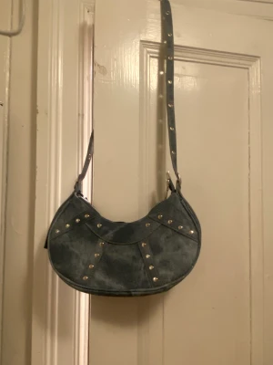 En blå axelbandväska - En blå axebandväska med metal detaljer på. Minns ej var den är ifrån. Den kommer inte till användning eftersom jag ej använder den.