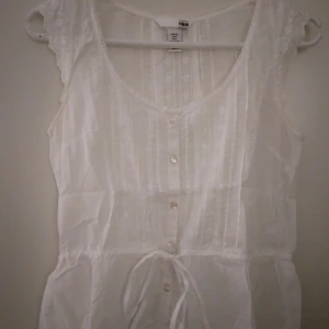 Vit romantisk ärmlös blus, vintage look. Strlk: 34, XS. Bomull. - Vit romantisk ärmlös blus/linne med knyt i midjan och broderier, vintage look. Strlk: 34, XS. Bomull. Ok skick, se bilder. Inget trasigt.