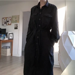 Svart klänning - Ginatricot - Svart klänning i längre modell, går ner till vaderna på mig som är 170. Jeansliknande men är i ett mjukt material (lyocell) som är väldigt luftigt och bekvämt, perfekt för sommarhalvåret! Två framfickor och knappar som gar hela vägen ned. Skärp ingår☺️