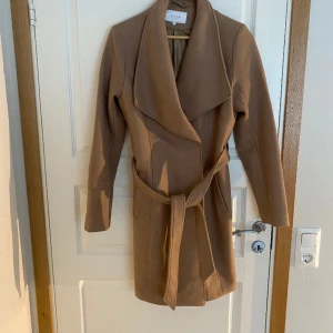 brun Kappa - En  beige/ brun kappa ifrån Märket Vila med 2 knappar och ett skärp. Använd 1 gång. 