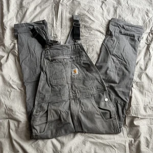Carhartt Hängselsbyxor - Carhart Duck Bib Stretch i grått.  Storlekt 32x32. Lite patina här och där!