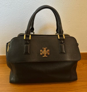 Tory Burch Mercer Dome Bag - Väskan är i fint skick förutom lite fläckar på insidan, har försökt tvätta bort de men har inte lyckats. Den har många olika fack och är rymlig.   Höjd: 24 cm Bredd: 32,5 cm Djup: 16 cm Handremmar: 12 cm