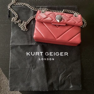 Kurt geiger - En snygg Kurt Gegier väska i liten modell. Lätt att bära och är den perfekta väskan om man ska gå bort.  Röd färg i skinn. Foder textil. Magnetlås. Detaljer är prydnadsstenar. Höjd: 14 cm Längd: 20 cm Vidd: 7 cm