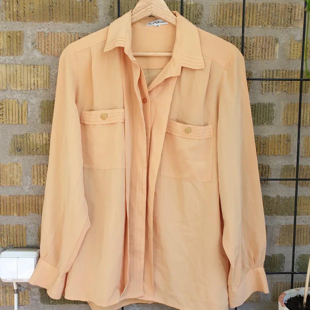 Tunn skjorta/blus i ett siden-liknande tyg, gul/orange/beige i färgen. Har många fina detaljer på kragen, fickorna och ärmarna och en sorts flärp över knapparna. Skulle kalle den för storlek M. Paidat.