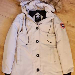 Canada Goose jacka (ej äkta) i ljusbeige med äkta päls på huvan. Mycket tjock och varm jacka med fina detaljer. Canada Goose dustbag och diverse informationslappar medföljer (som kommit med jackan). Skickar gärna fler bilder. 