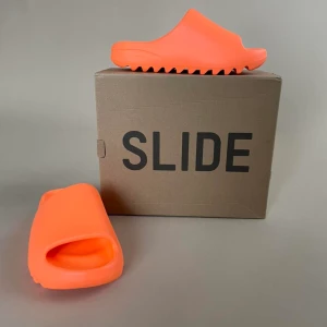 Brand New Yeezy x Adidas SlidesOriginal Brand - Brand New, Never Worn. Make a bold statement with these super comfy slides Color: Enflamed Orange US/UK Size 5  It’s recommended by retailers to go a full size up from your usual size.  Made of Resin Comes with Original packaging