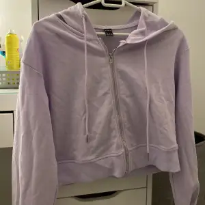 En lila ganska kort tröja med dragkedja, snörning & luva. Baktill har den ett rosa tryck med text på. Helt oanvänd och super fin. Skönt material. 40kr+frakt