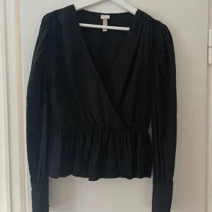 Svart blus med volang  - Jättefin svart blus från HM. Passar storlek 36/38. Puffärm och volang i midjan, v-ringning med en liten hake som håller ihop tröjan framtill.
