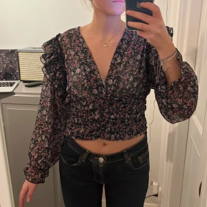 Blommig blus - Blommig croppad blus. Dragkedja i sidan. Volanger på axlarna och i slutet på ärmarna!