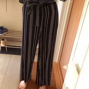 Långbyxor stl. 36 - Randiga långbyxor i storlek 36 från Na-kd. Svarta led vita asymmetriska ränder. Ankellånga och hög midja med dekorativt band. Fickor i sidan. 100% viskos, skönt material. Fint skick, använda endast få ggr.   Skriv för fler bilder! 