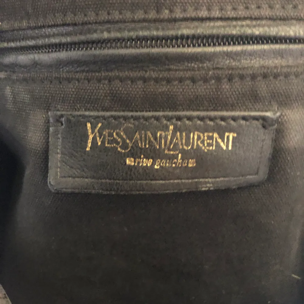 Säljer min väska från Yves Saint Laurent. Den är använd men i fint skick. Den är den perfekta rymliga vardagsväskan som rymmer mycket. En axelväska från YSL är alltid trendigt. Svart skinn och spännen i mässing. Äkta med märke inuti och broderat i botten.. Väskor.