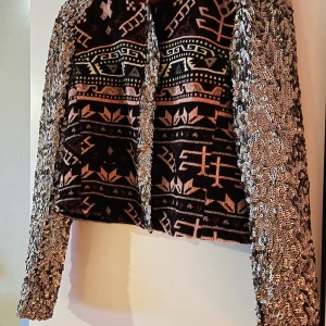 Kort jacka/top med paljetter - Har använts både som jacka och festtopp.  Tyget är mönstrat i vinröda och rosa toner och ärmarna är täckta i silvriga paljetter.  Storlek M men liten i storleken, ”modellen på bild” är en medium och den sitter tajt.  Köparen står för frakten.  