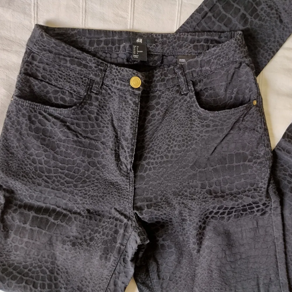 Snygga svarta/mörkgrå högmidjade byxor med guldfärgade detaljer från H&M. Sytt snakeskininspirerat mönster. Tyget är pyttelite nopprigt/ludet på vissa ställen. 55% viskos 41% bomull 4% elastan. Storlek 36. . Jeans & Byxor.