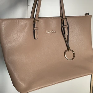 Väska - Bege handväska med guld detaljer, använd som skolväska i grundskolan men inte använd på ungefär 5 år, bra skick förutom det svarta märket på undersidan. Minns inte vart jag köpt den. Den är på ett ungefär 28x34 försökte mäta med linjal.