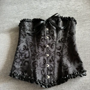 Svart korsett med mönster - En elegant svart korsett med ett vackert mönster. Korsetten har knappar framtill och snörning baktill för justerbar passform. Den är dekorerad med volanger längs över- och nederkanten samt en rosett framtill.