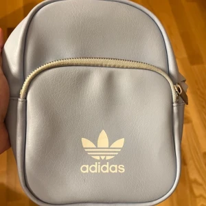 Ljusblå axelväska från Adidas - En ljusblå axelväska från Adidas med dragkedja och justerbar axelrem. Väskan har en framficka med dragkedja och Adidas-logotypen tryckt på framsidan.