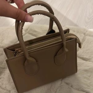 Beige handväska - En elegant beige handväska med två handtag och dragkedja. Väskan har en kompakt design med flera fack inuti för organisering.