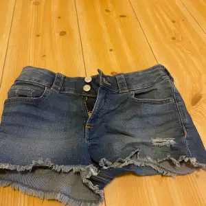 Ett par blå jeansshorts med fransade kanter och slitna detaljer. Shortsen har två framfickor, två bakfickor, och stängs med dragkedja och två knappar.