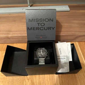 Omega x Swatch Mission to Mercury - En snygg klocka från Omega x Swatch Bioceramic Moonswatch Collection, modell Mission to Mercury. Klockan har en svart urtavla med vita detaljer och en grå rem. original box och alla papper finns inklusive kvitto. Pris på merchsweden: 6000 kronor 