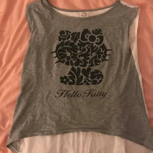 Grå Hello Kitty topp - En grå ärmlös topp från Hello Kitty med ett svart mönster av Hello Kitty-figuren på framsidan. Toppen har en rund halsringning och är tillverkad i ett mjukt material.