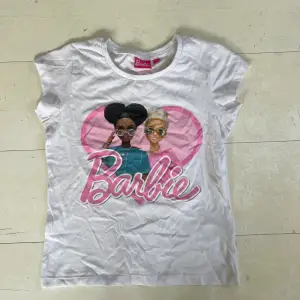 En vit t-shirt från Barbie med ett tryck av två Barbie-figurer och ett rosa hjärta i bakgrunden. T-shirten har korta ärmar och en rund halsringning. passar någon med storlek XS.