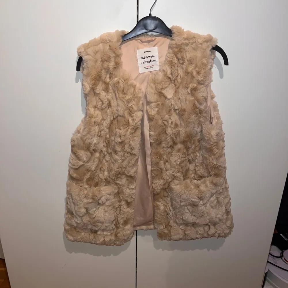 En mjuk och fluffig beige pälsväst från Zara's Outerwear Collection. Västen har en öppen design utan knappar eller dragkedja och är fodrad med ett slätt tyg på insidan. västen passar även storleken Xs/S. Jackor.