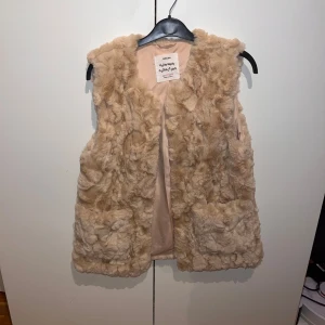 Beige pälsväst från Zara - En mjuk och fluffig beige pälsväst från Zara's Outerwear Collection. Västen har en öppen design utan knappar eller dragkedja och är fodrad med ett slätt tyg på insidan. västen passar även storleken Xs/S