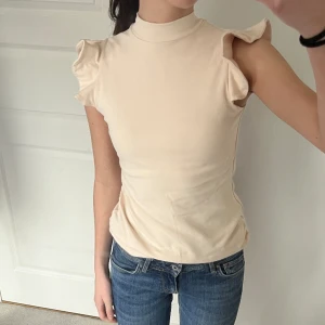 Söt beige topp - Söta volanger på ärmarna och liten krage. Sitter tajt på och är i tjockt material. Använd typ en gång så toppen skick! 