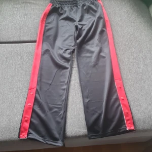 Träningsbyxor - MLY träningsbyxor/fritidsbyxa med gummiband i midjan. Röda ränder på sidorna, med 4 knappar vid varje vad. 