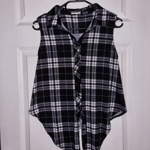 Svart och vit rutig ärmlös skjorta - En svart och vit rutig ärmlös skjorta med knappar framtill. Skjortan har en klassisk krage och en lös passform.