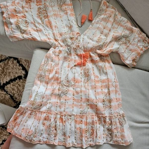 Sommarklänning i viskos - En vacker klänning med ett vackert mönster i aprikosfärg och krämfärgad med gulddetaljer. Klänningen har en V-ringning och korta ärmar. Den har en snörning i midjan och en volang i nederkanten. Viskos 100% Aldrig använd.Helt ny!